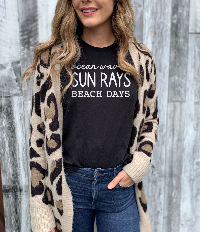 May include: A woman wearing a black t-shirt with the text "ocean waves sun rays beach days" printed on it. She is wearing a brown and white leopard print cardigan and blue jeans.