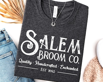 May include: A dark grey heather t-shirt featuring white distressed text that reads SALEM BROOM CO. Quality Handcrafted Enchanted EST 1692. The shirt has a crew neckline and is displayed with a silver sunburst pendant necklace resting on the fabric.
