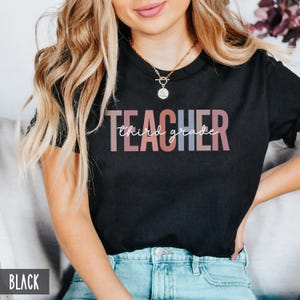 A woman wearing a black t-shirt featuring pastel block letters spelling teacher and white cursive text that reads third grade.