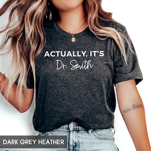 May include: A dark grey heather t-shirt with the text "Actually, it's Dr. Smith" printed in white.