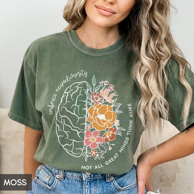 以下が含まれることがあります： 片側に花がついた脳の白い輪郭が描かれた緑色のTシャツ。Tシャツには「Embrace Neurodiversity Not All Great Minds Think Alike」という文字が印刷されています。