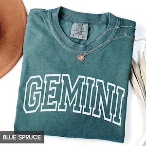 May include: Blue spruce t-shirt with the word "GEMINI" in white block letters. The shirt is folded and has a gold necklace with a pendant. The brand label "Comfort Colors" is visible. A brown hat is partially visible in the background.