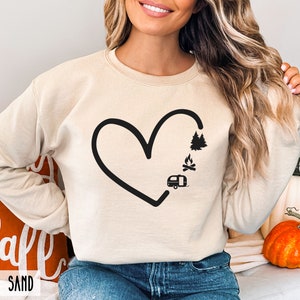 May include: A cream-colored sweatshirt with a black outline of a heart. Inside the heart are black silhouettes of a camper, a campfire, and a pine tree.