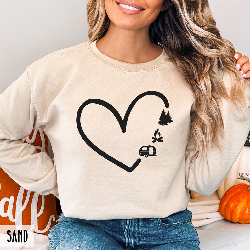 May include: A cream-colored sweatshirt with a black outline of a heart. Inside the heart are black silhouettes of a camper, a campfire, and a pine tree.