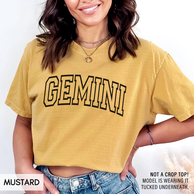 May include: Mustard-colored t-shirt with the word "GEMINI" in black block letters. The shirt has a vintage, slightly faded look. The text "NOT A CROP TOP! MODEL IS WEARING IT TUCKED UNDERNEATH." is also visible.