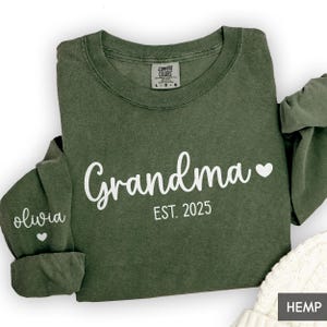 May include: An olive green long-sleeve crewneck sweatshirt featuring white text that reads Grandma EST. 2025 with a heart icon. The sleeve displays the name olivia with a small heart. A Comfort Colors brand label is visible at the neck.