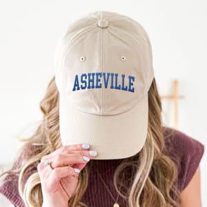Puede incluir: Gorra de b&eacute;isbol beige con la palabra "ASHEVILLE" bordada en azul en la parte delantera. La gorra tiene una visera curva y un bot&oacute;n en la parte superior. La gorra est&aacute; siendo sostenida por una persona.