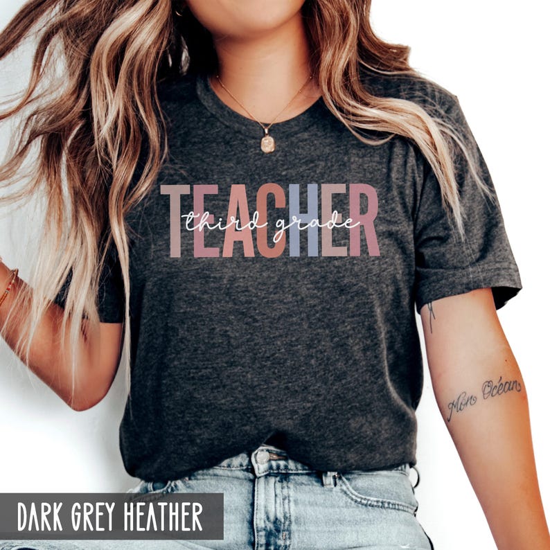 A woman wearing a dark grey heather t-shirt printed with block letters spelling teacher and white cursive text spelling third grade.