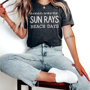 May include: A dark gray t-shirt with white text that reads "ocean waves SUN RAYS BEACH DAYS". The model is wearing the t-shirt with light blue jeans and white sneakers.