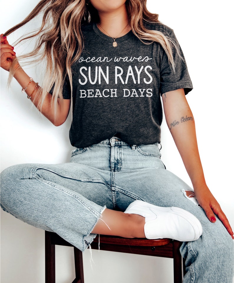 May include: A dark gray t-shirt with white text that reads "ocean waves SUN RAYS BEACH DAYS". The model is wearing the t-shirt with light blue jeans and white sneakers.