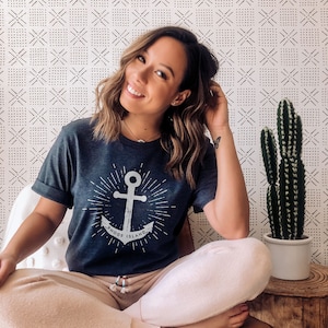 May include: A woman wearing a dark blue t-shirt with a white anchor graphic and the text "Rhode Island". She is sitting on a brown chair with her legs crossed and wearing light pink sweatpants.