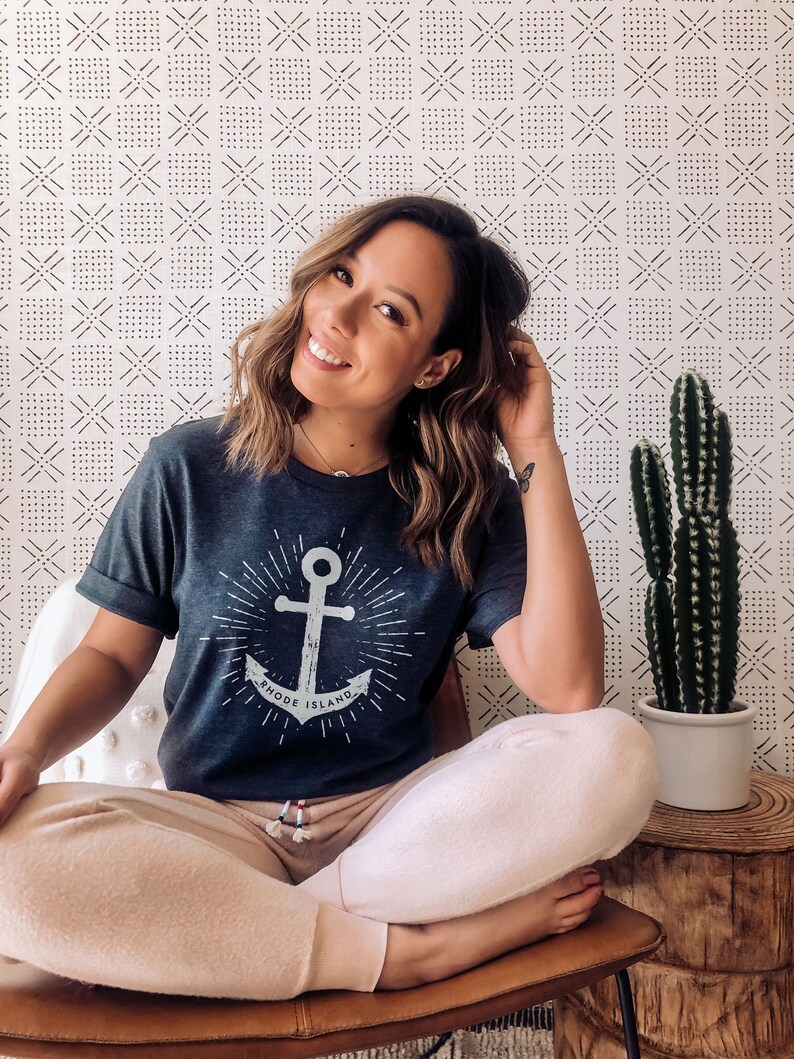 May include: A woman wearing a dark blue t-shirt with a white anchor graphic and the text "Rhode Island". She is sitting on a brown chair with her legs crossed and wearing light pink sweatpants.