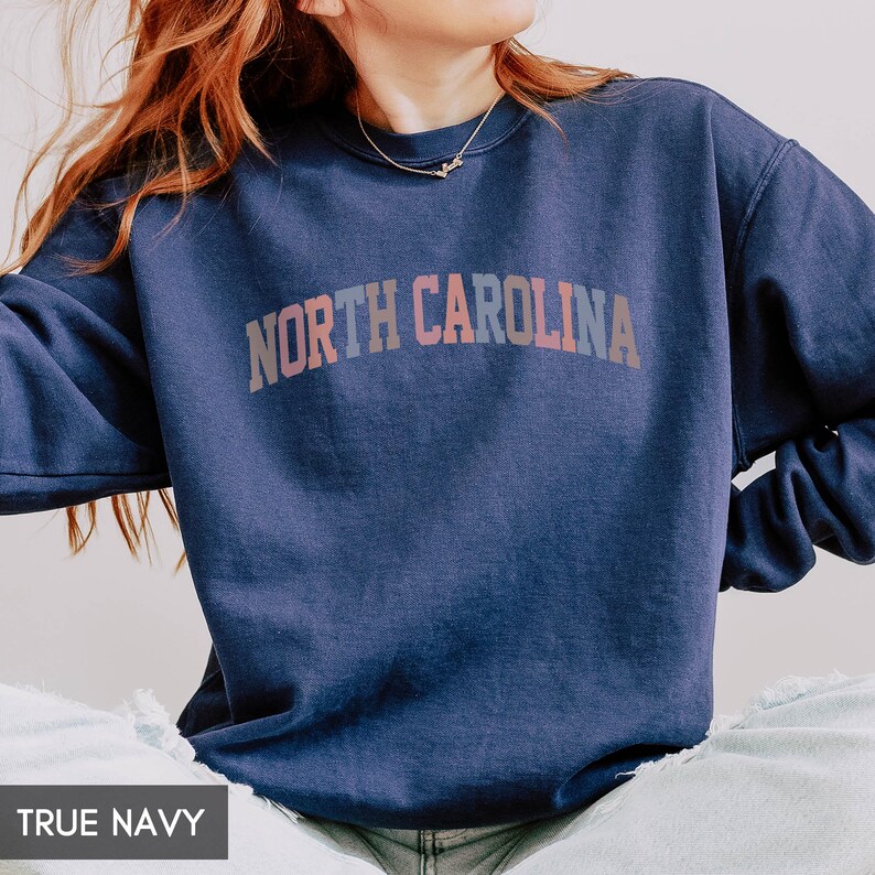 Puede incluir: Una sudadera azul marino con el texto "NORTH CAROLINA" en una fuente arqueada multicolor. La sudadera es de color liso y parece estar hecha de un material suave. Las palabras "TRUE NAVY" est&aacute;n en la parte inferior de la imagen.