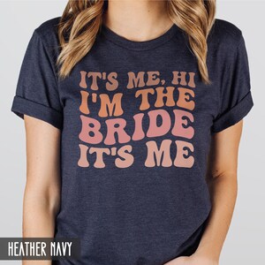 May include: A heather navy blue t-shirt with the text "It's me, hi I'm the bride it's me" in a retro font with a gradient of pink and orange.