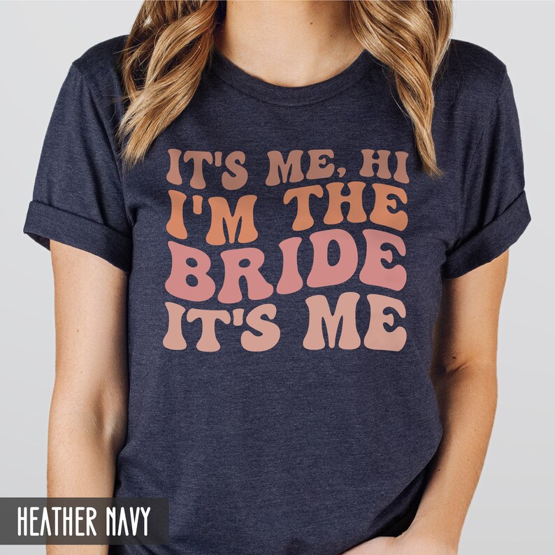 May include: A heather navy blue t-shirt with the text "It's me, hi I'm the bride it's me" in a retro font with a gradient of pink and orange.