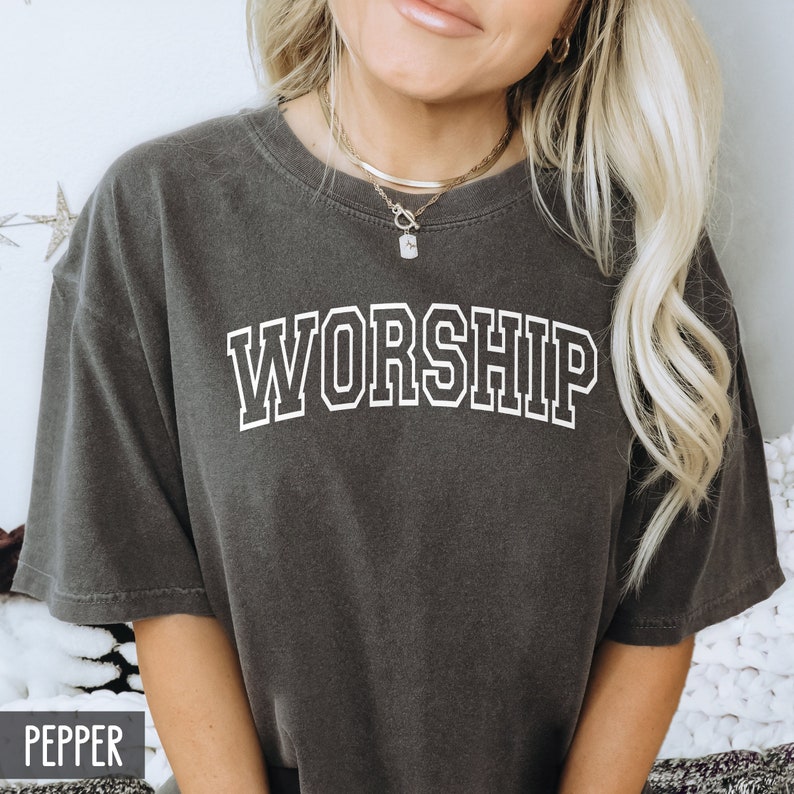 K&ouml;nnte beinhalten: Ein dunkelgraues T-Shirt mit dem Wort "WORSHIP" in Wei&szlig; mit wei&szlig;em Umriss gedruckt. Das Shirt wird von einer Person mit langen blonden Haaren getragen.
