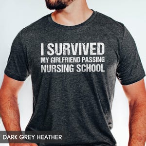 May include: A dark grey heather short-sleeve t-shirt featuring white distressed text that reads I SURVIVED MY GIRLFRIEND PASSING NURSING SCHOOL. The shirt has a crew neckline and a textured fabric appearance with a visible blend of dark and light fibers.