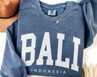 Peut inclure: Sweat-shirt bleu denim avec le mot "BALI" en grandes lettres blanches et "INDONESIA" en dessous. Un collier en or avec un pendentif est drapé sur le sweat-shirt. Un chapeau marron est visible en arrière-plan.
