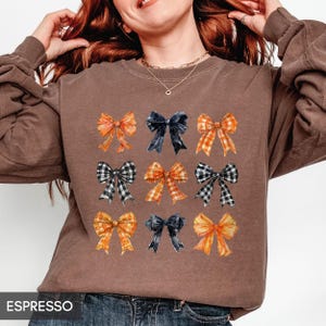 May include: Espresso-colored sweatshirt featuring nine watercolor bows in various patterns and colors, including orange, navy, and black and white gingham. The word "ESPRESSO" is printed at the bottom of the image. The bows are arranged in three rows of three.