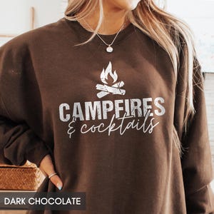 May include: Dark chocolate brown sweatshirt with the text "Campfires & Cocktails" in white, along with a graphic of a campfire. The sweatshirt is a crew neck style.