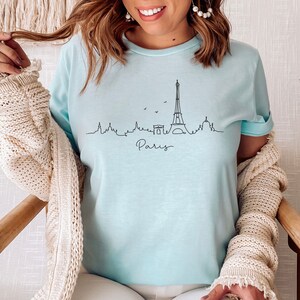 Op de afbeelding: Een lichtblauw t-shirt met een zwarte omtrek van de Eiffeltoren en het woord "Paris" eronder.