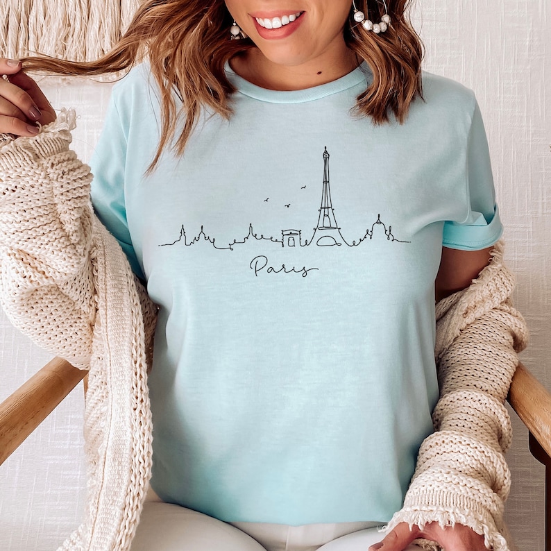 Op de afbeelding: Een lichtblauw t-shirt met een zwarte omtrek van de Eiffeltoren en het woord "Paris" eronder.