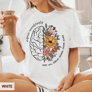 以下が含まれることがあります： 脳に花が描かれた白いTシャツ。片側に「Embrace neurodiversity. Not all great minds think alike.」というテキストが書かれています。