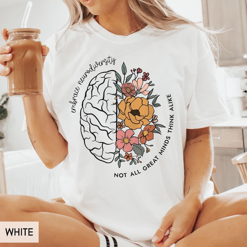 以下が含まれることがあります： 脳に花が描かれた白いTシャツ。片側に「Embrace neurodiversity. Not all great minds think alike.」というテキストが書かれています。