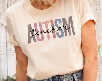 May include: A soft cream short-sleeve t-shirt featuring the word AUTISM in large, vertical, multi-colored block letters with the word teacher written in a black cursive script font layered horizontally across the center of the design.