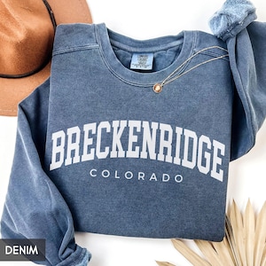 May include: A denim blue sweatshirt with the word "BRECKENRIDGE" in white block letters, with "COLORADO" below. A gold necklace with a pendant and a brown hat are visible. The sweatshirt is a Comfort Colors brand.