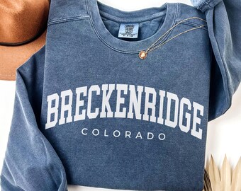 Peut inclure: Un sweat-shirt bleu denim avec le mot "BRECKENRIDGE" en lettres blanches, avec "COLORADO" en dessous. Un collier doré avec un pendentif et un chapeau marron sont visibles. Le sweat-shirt est de la marque Comfort Colors.