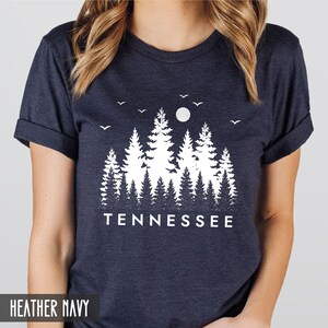 May include: A heather navy blue t-shirt with a white graphic design of a forest silhouette with a moon and birds. The text "TENNESSEE" is printed below the graphic.