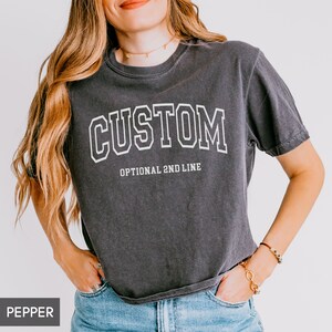 May include: A dark gray cropped t-shirt with the word "CUSTOM" in white block letters and the text "OPTIONAL 2ND LINE" in smaller white letters below. The shirt is being worn by a person with long brown hair.