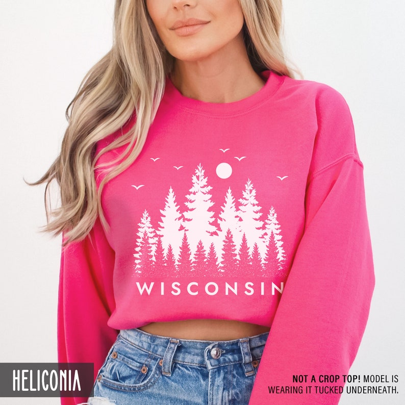 May include: Pink sweatshirt with a white graphic of trees and the word "WISCONSIN". The model is wearing the sweatshirt tucked into blue jeans.