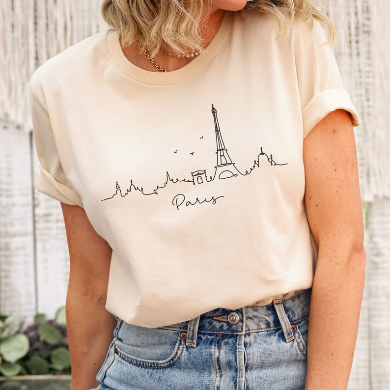 Op de afbeelding: Een cr&egrave;mekleurig T-shirt met een zwarte lijntekening van de Eiffeltoren en het woord "Paris" in cursief.
