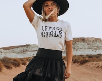 Peut inclure: Un t-shirt blanc à manches courtes avec le texte noir LET'S GO GIRLS imprimé sur le devant, associé à un chapeau noir à larges bords et une jupe noire taille haute avec un ourlet à volants et une large ceinture.