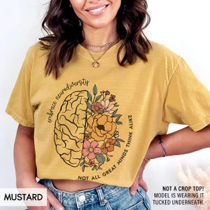 以下が含まれることがあります： マスタードイエローのTシャツに、花が咲いた脳のグラフィックと「embrace neurodiversity not all great minds think alike」というテキストがプリントされています。