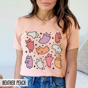 Op de afbeelding: Een perzikkleurig T-shirt met een grafische print van kleurrijke katten en geesten. Het shirt heeft een ronde hals en korte mouwen. De tekst "HEATHER PEACH" is gedrukt op de onderkant van het shirt.