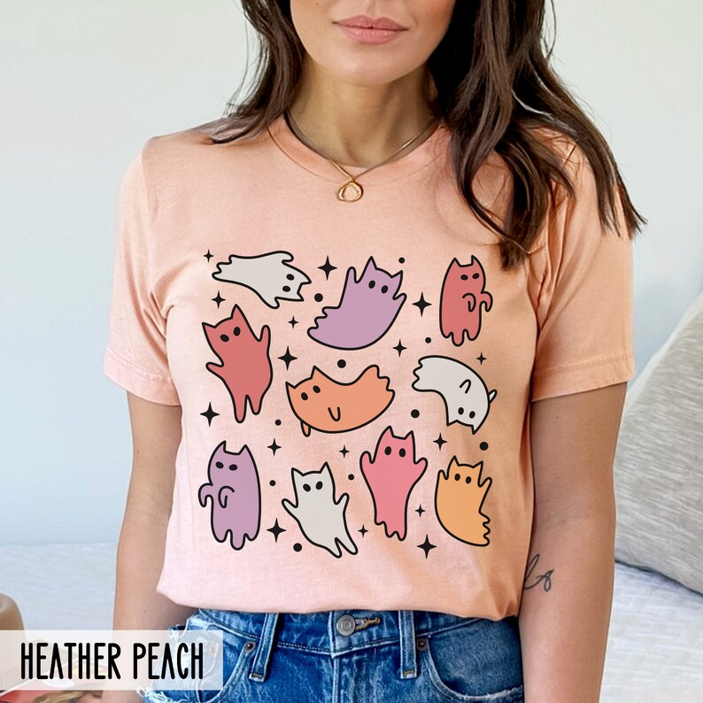Op de afbeelding: Een perzikkleurig T-shirt met een grafische print van kleurrijke katten en geesten. Het shirt heeft een ronde hals en korte mouwen. De tekst "HEATHER PEACH" is gedrukt op de onderkant van het shirt.
