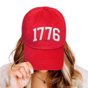 May include: A red baseball cap with the year "1776" embroidered in white block letters. The hat is a classic six-panel design with a curved brim. The cap is made of a soft, durable material.