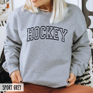 A person wearing a sport grey crewneck sweatshirt with the word HOCKEY in black collegiate block letters.