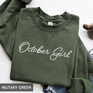 May include: A military green long-sleeve sweatshirt featuring the text October Girl printed in white cursive script across the chest. The garment has a crew neckline and a ribbed collar, cuffs, and hem, showcasing a solid dark green color.