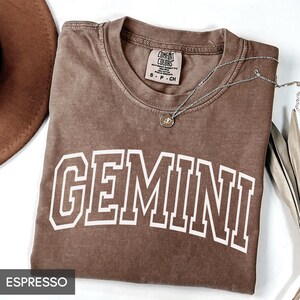 May include: Espresso-colored t-shirt with the word "GEMINI" in white block letters. A silver necklace with a pendant rests on the shirt. The shirt is folded, and a brown hat is partially visible in the upper left corner.
