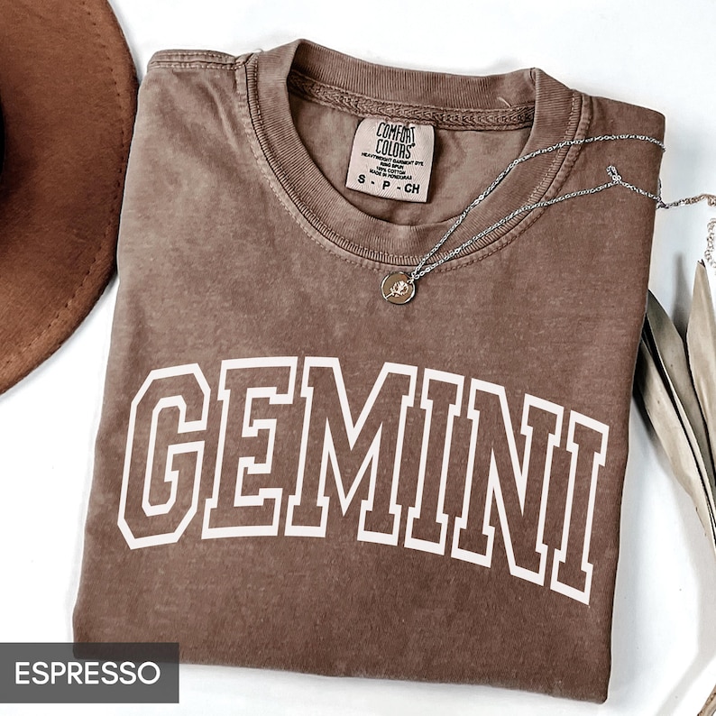 May include: Espresso-colored t-shirt with the word "GEMINI" in white block letters. A silver necklace with a pendant rests on the shirt. The shirt is folded, and a brown hat is partially visible in the upper left corner.