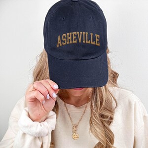 Puede incluir: Gorra de b&eacute;isbol azul marino con la palabra "ASHEVILLE" bordada en hilo dorado. La gorra es de estilo cl&aacute;sico con una visera curva y una correa ajustable. Una persona sostiene la gorra.