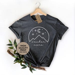 A dark gray t-shirt hanging on a wooden hanger features a white graphic of mountains, a crescent moon, and stars with the text wander more.