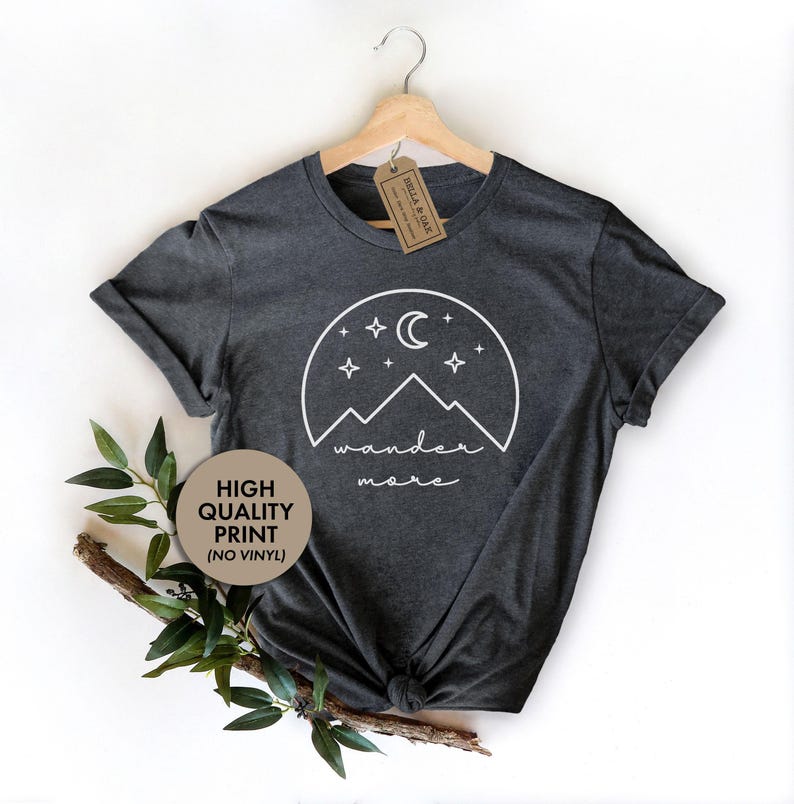 A dark gray t-shirt hanging on a wooden hanger features a white graphic of mountains, a crescent moon, and stars with the text wander more.