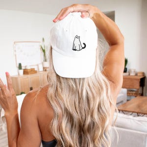 May include: White baseball cap with a black cat graphic. The cap is made of a soft material and has a curved brim. The cat graphic is a simple, minimalist design. The cap is being held up by a person.