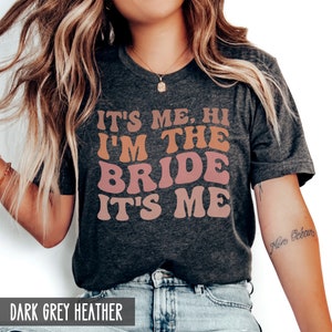 May include: A dark grey heather t-shirt with the text "It's me, hi I'm the bride it's me" in a retro font with orange, pink, and red colors.