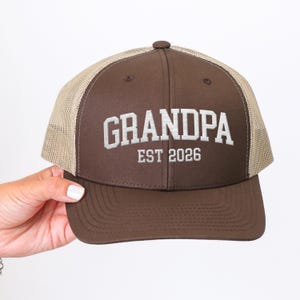 May include: A brown trucker hat with a mesh back and a curved brim. The front of the hat features the embroidered word "GRANDPA" above "EST 2026" in white block letters. The hat is being held by a person.